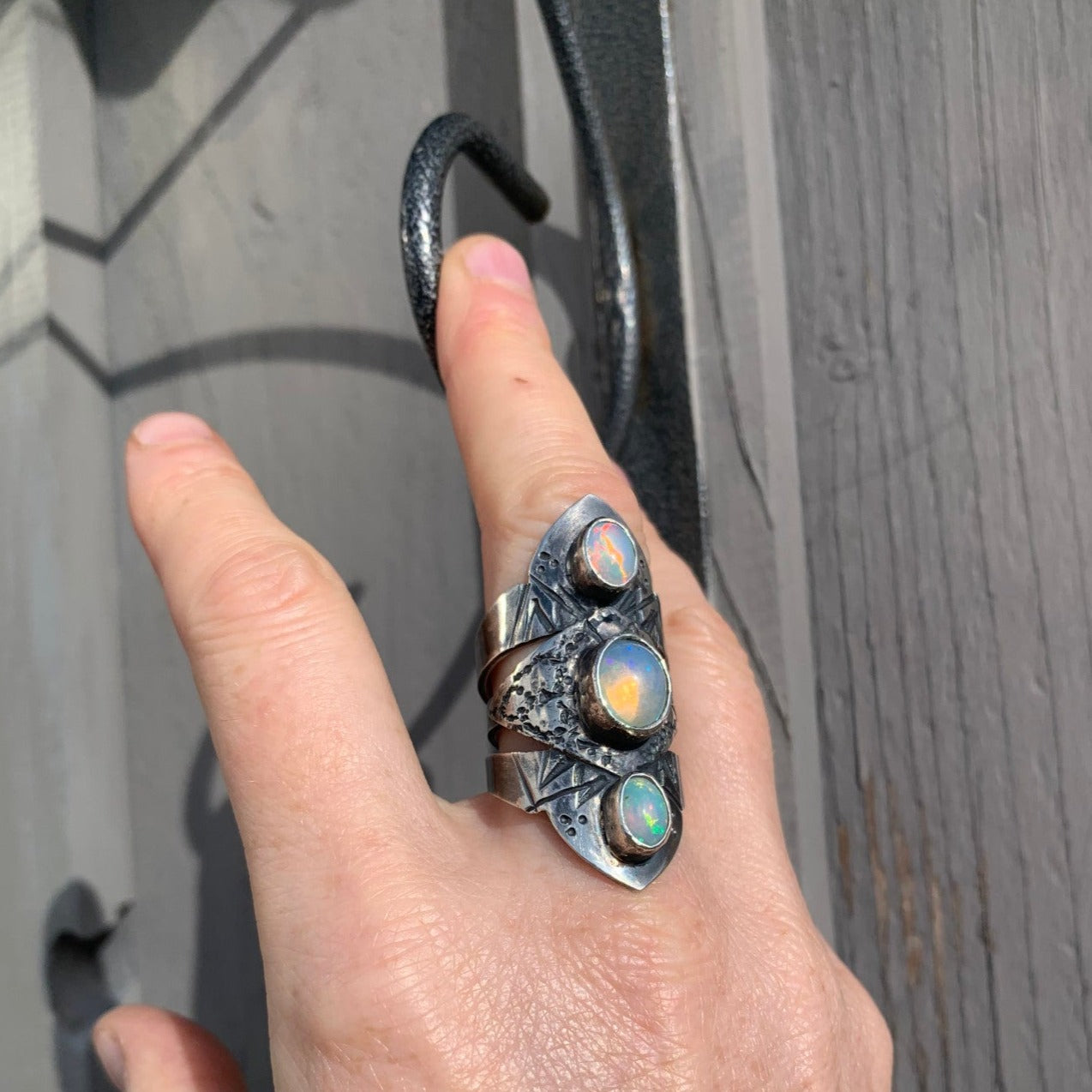 Bague composée de 3 joncs soudés et gravés de symboles berbères (serpent lunaire, grains et gâteau de miel), sertie de trois opales Welo éthiopiennes. Le tout représentent la lune en harmonie avec le serpent lunaire, illustrant une syzygie – un moment où trois corps célestes sont alignés, reliant les cycles de la lune et de la mue du serpent. Opales laiteuses aux reflets rose, jaune et bleu selon l'orientation du soleil. Vue du dessus portée à l'annulaire droit.