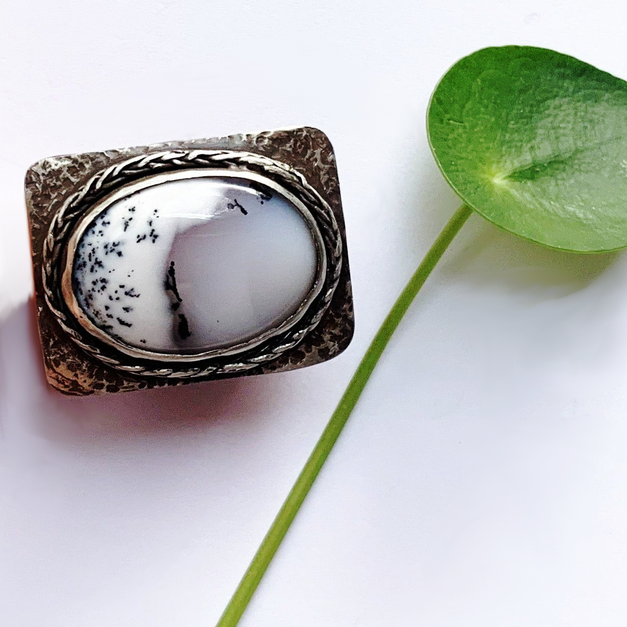 Grosse bague en argent martelé présentant un plateau rectangulaire aux coins arrondis, sertis d'une incroyable Agathe dendritique d'un blanc laiteux avec des veines noires d'oxyde de manganèse  lui donnant un aspect de paysage sous la neige, le tour du serti est souligné d'un brin de tresse en argent soudé pour lier le sort. Le dessous du serti est évidé pour laisser passer la lumière dans la pierre. taille 65. vue de face