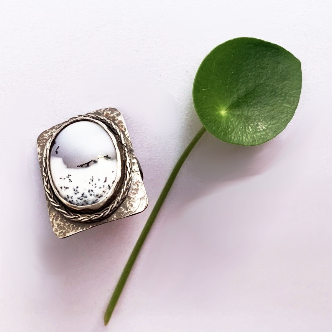 Grosse bague en argent martelé présentant un plateau rectangulaire aux coins arrondis, sertis d'une incroyable Agathe dendritique d'un blanc laiteux avec des veines noires d'oxyde de manganèse  lui donnant un aspect de paysage sous la neige, le tour du serti est souligné d'un brin de tresse en argent soudé pour lier le sort. Le dessous du serti est évidé pour laisser passer la lumière dans la pierre. taille 65. vue de profil