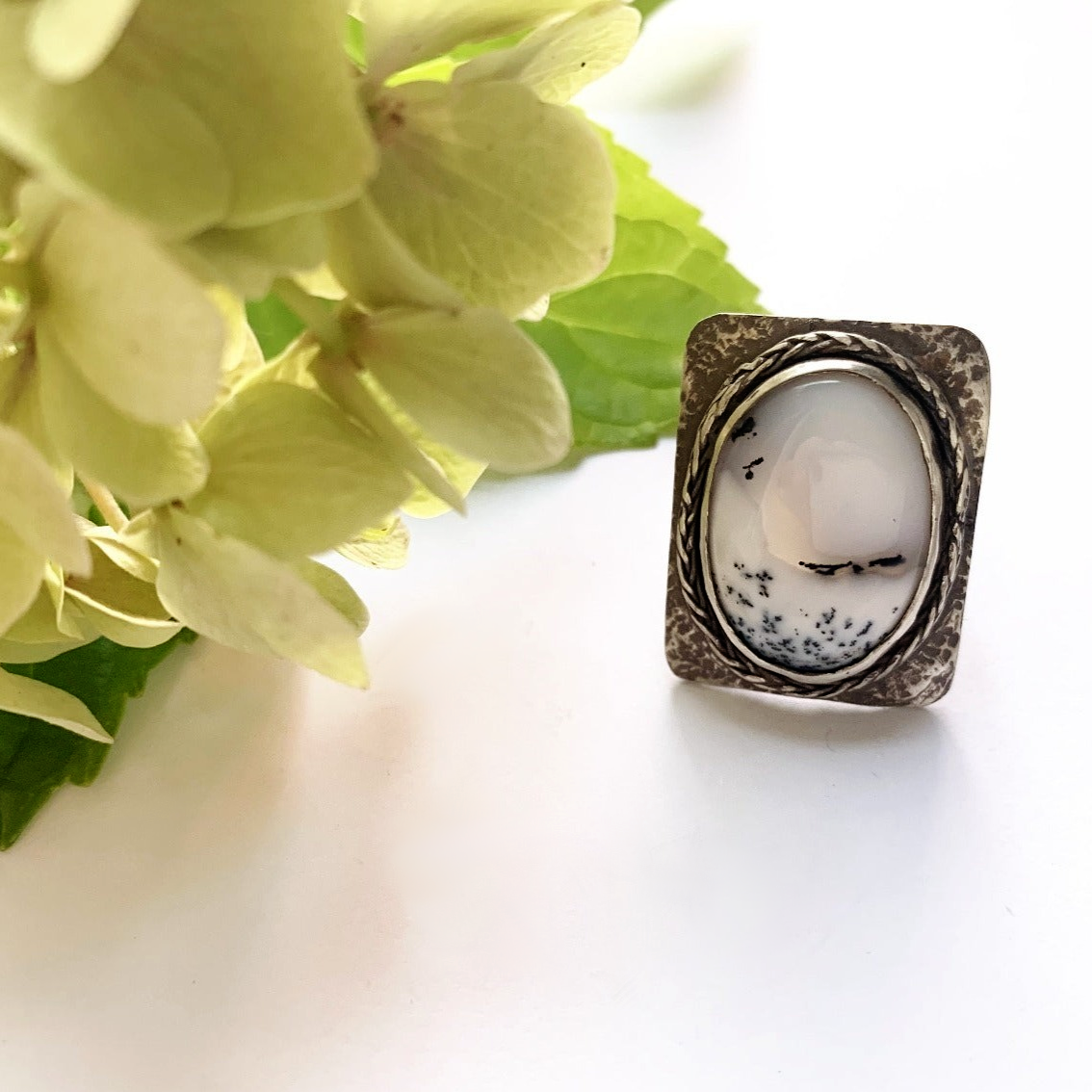 Grosse bague en argent martelé présentant un plateau rectangulaire aux coins arrondis, sertis d'une incroyable Agathe dendritique d'un blanc laiteux avec des veines noires d'oxyde de manganèse  lui donnant un aspect de paysage sous la neige, le tour du serti est souligné d'un brin de tresse en argent soudé pour lier le sort. Le dessous du serti est évidé pour laisser passer la lumière dans la pierre. taille 65. vue de face