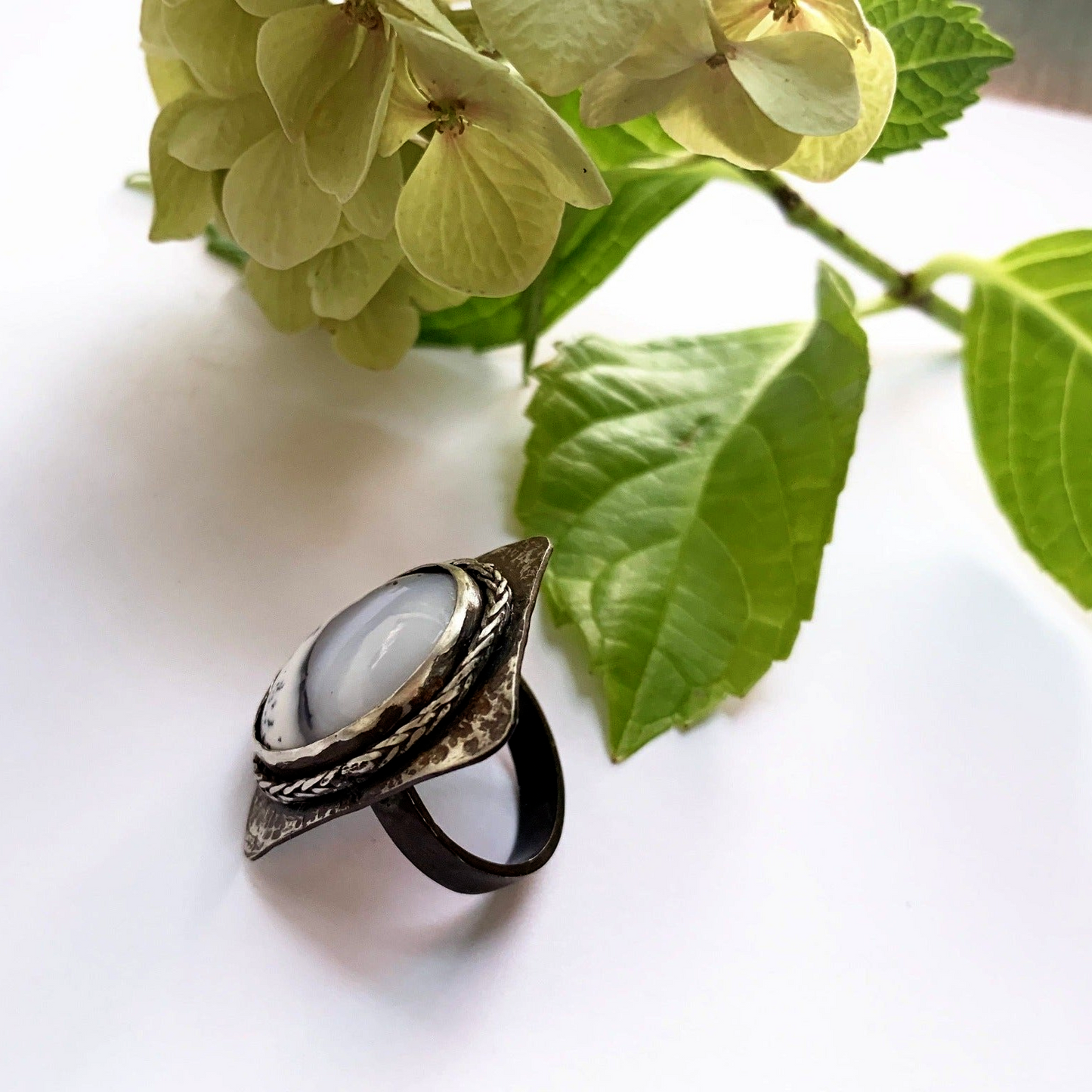 Grosse bague en argent martelé présentant un plateau rectangulaire aux coins arrondis, sertis d'une incroyable Agathe dendritique d'un blanc laiteux avec des veines noires d'oxyde de manganèse  lui donnant un aspect de paysage sous la neige, le tour du serti est souligné d'un brin de tresse en argent soudé pour lier le sort. Le dessous du serti est évidé pour laisser passer la lumière dans la pierre. taille 65. vue de coupe