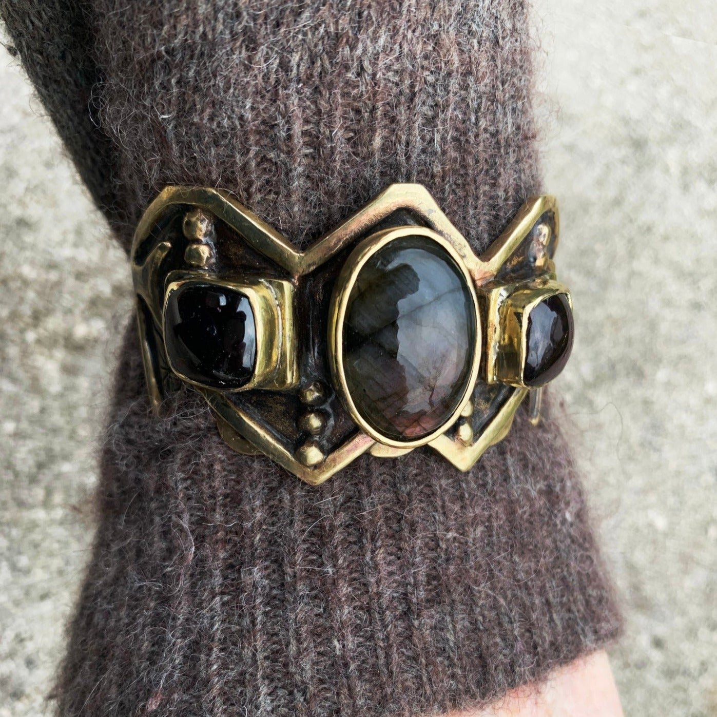 Bracelet de fonte en laiton, sertis d'une labradorite et de deux grenats. Le haut est découpé en W, symbole berbère de la fourdre, et le bas en M (symbole de fécondité), sous chaque serti il y a un boitier ouvert contenant un symbole berbère. Le bracelet est lourd 80 grammes et prévu pour un poignet de 18 cm avec ouverture de 4 cm agrandissable sur demande. Vue porté. Patine noircissante, et verni. 