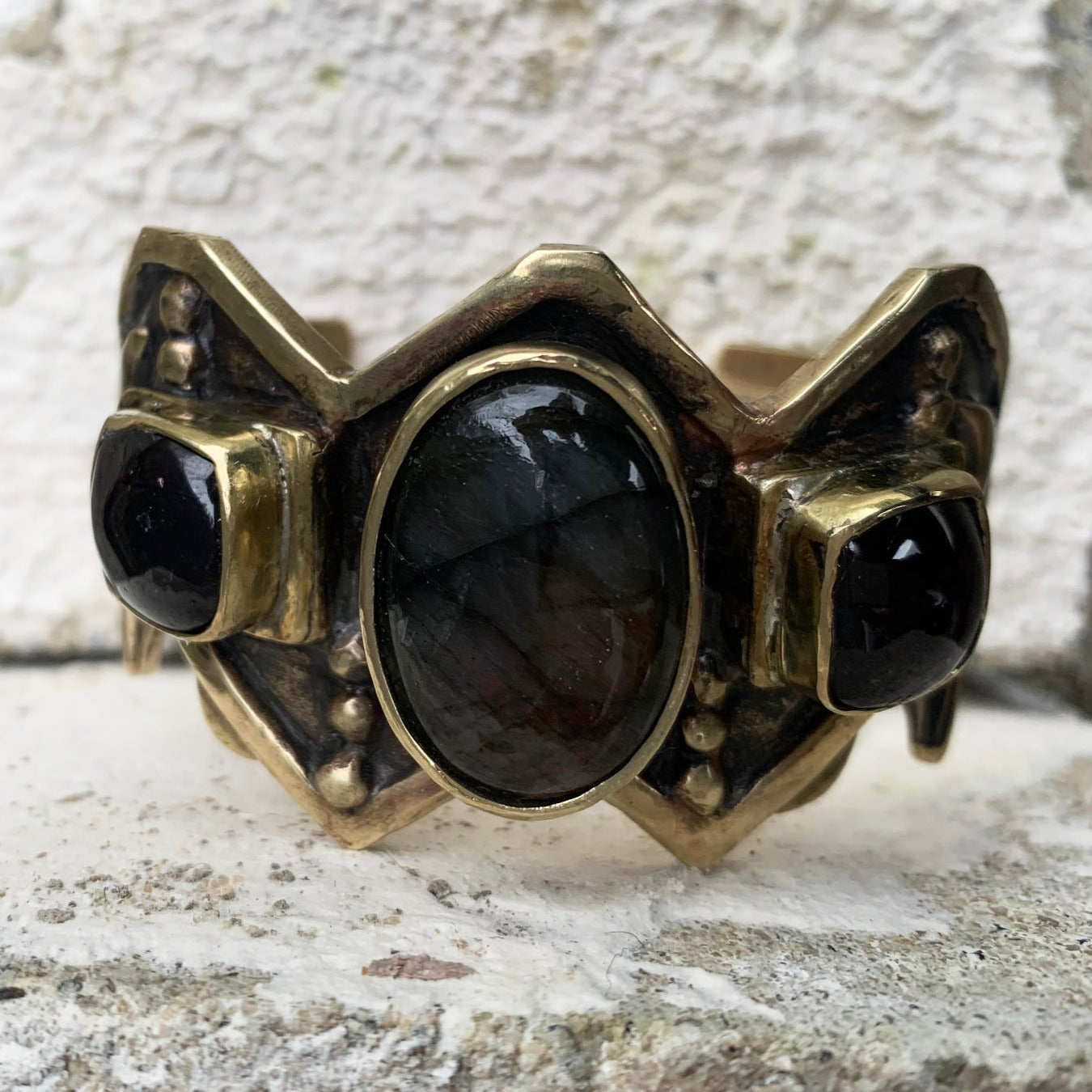 Bracelet de fonte en laiton, sertis d'une labradorite et de deux grenats. Le haut est découpé en W, symbole berbère de la fourdre, et le bas en M (symbole de fécondité), sous chaque serti il y a un boitier ouvert contenant un symbole berbère. Le bracelet est lourd 80 grammes et prévu pour un poignet de 18 cm avec ouverture de 4 cm agrandissable sur demande. Vue en de face. Patine noircissante, et verni. 