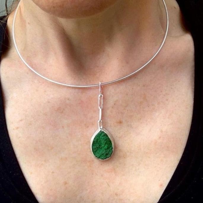 Et si vous accrochiez un fragment de roche à votre cou ? Ce collier en argent de 40 cm présente au centre une uvarovite druzy sertie clos pour la protéger des chocs. Sa forme druzy fait qu'elle est recouverte de minuscules inclusions brillants au soleil. Ce pendentif est long de 5 cm, accroché par des chaînons rectangles en argent. Vue en gros plan du serti-clos de face avec la druze qui semble déborder du serti par ses reliefs naturels.