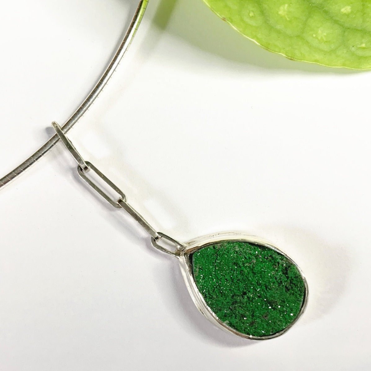 Et si vous accrochiez un fragment de roche à votre cou ? Ce collier en argent de 40 cm présente au centre une uvarovite druzy sertie clos pour la protéger des chocs. Sa forme druzy fait qu'elle est recouverte de minuscules inclusions brillants au soleil. Ce pendentif est long de 5 cm, accroché par des chaînons rectangles en argent. Vue en gros plan du serti-clos de face avec la druze qui semble déborder du serti par ses reliefs naturels.