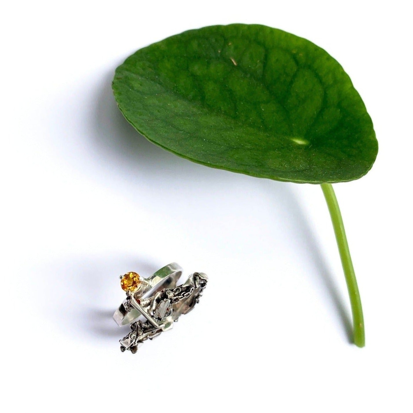 Inspirée par les couronnes de fleurs de Tahiti, cette bague évoque le Lei, symbole de l'Aloha hawaïen. Elle se compose de deux joncs : un petit en taille 62 avec une citrine de 5 mm, porté côté main, et un jonc plus large pour le pouce, constitué de morceaux d'argent travaillés. Les deux joncs sont reliés par un pont en forme de tige de plante, visible de dessus coté citrine.