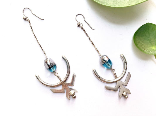 Boucles d'oreilles en argent composées d'un crochet ouvert, sur lequel est suspendue une chaîne en maille forçat en argent de 3 cm. Sur cette chaîne est glissée une perle de verre bleu translucide, dont un côté a été argenté en surface. Sous cette perle est accroché un demi arc marqué de points du foyer, symbolisant la nouvelle lune et le changement de cap dans la vie. Sur cet arc est soudé un symbole de femme en argent, les jambes écartées en W à l'envers, en position de donner la vie.