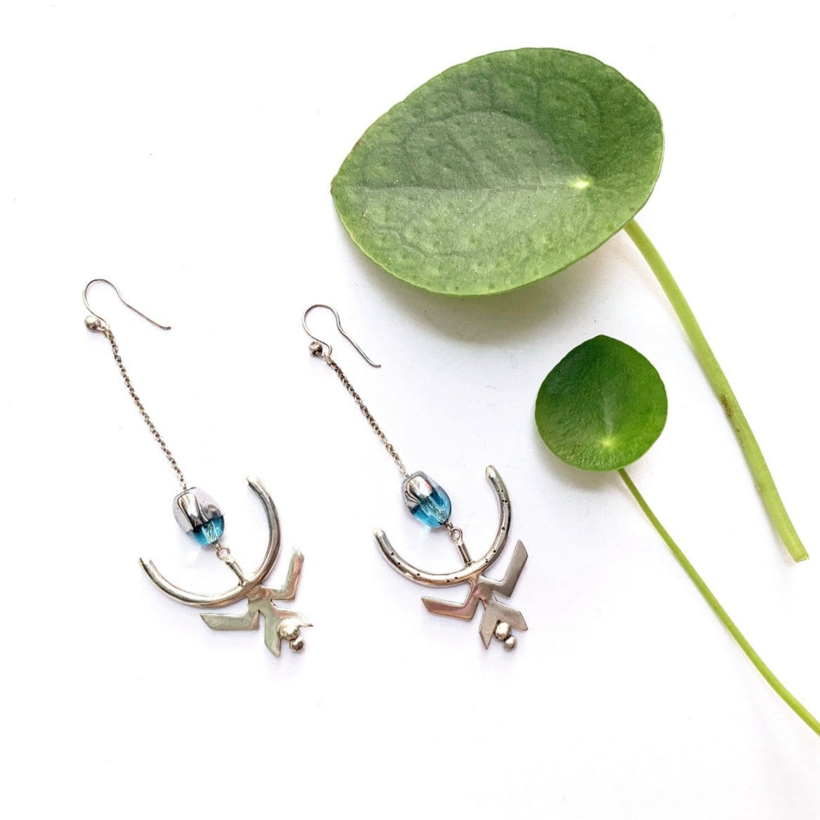 Boucles d'oreilles en argent composées d'un crochet ouvert, sur lequel est suspendue une chaîne en maille forçat en argent de 3 cm. Sur cette chaîne est glissée une perle de verre bleu translucide, dont un côté a été argenté en surface. Sous cette perle est accroché un demi-arc marqué de points du foyer, symbolisant la nouvelle lune et le changement de cap dans la vie. Sur cet arc est soudé un symbole de femme en argent, les jambes écartées en W à l'envers, en position de donner la vie. Vue éloignées.