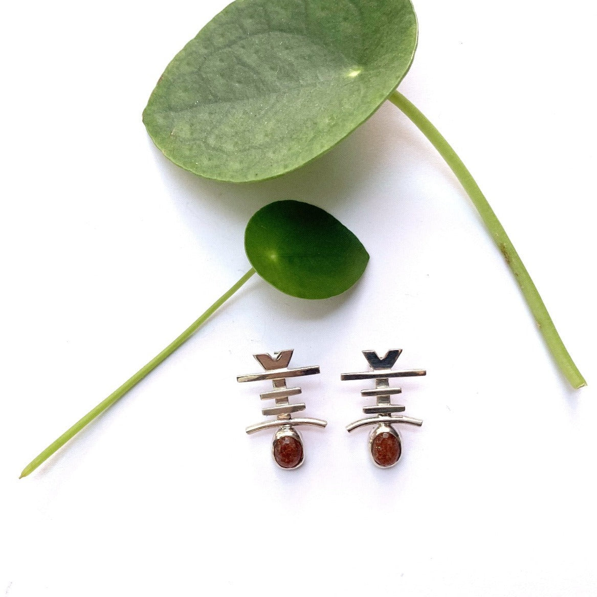 Petites boucles d'oreilles en argent, fixées par des tiges et des papillons pour oreilles percées. En forme de peigne berbère, certains diraient qu'elles ressemblent à de petits poissons. Elles sont modelées en forme de Y, sur lequel sont soudées trois barres d'argent de différentes tailles, puis un petit arc, sous lequel est sertie une magnifique pierre de soleil orangé, avec des éclats d'hématite qui brillent en captant la lumière