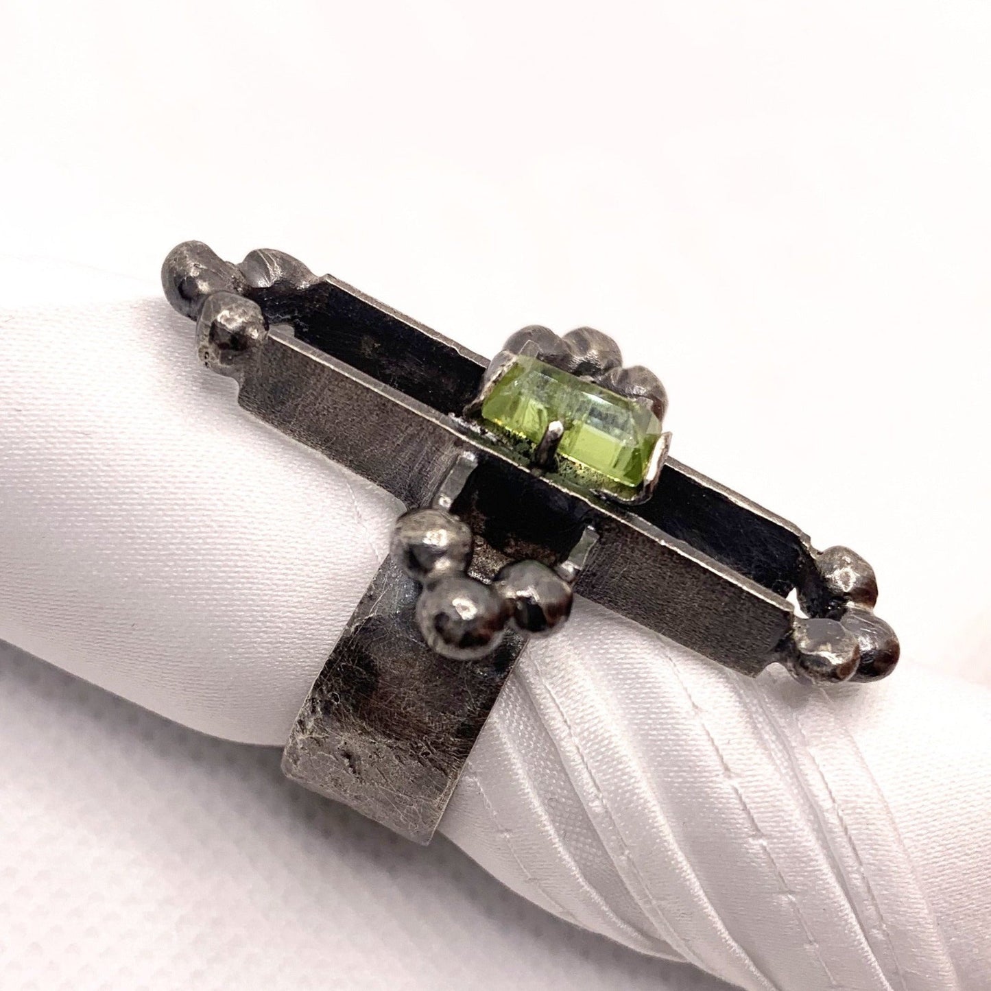 Bague en argent massive et texturé, sur le doigt un soleil amazigh soit une croix avec son centre déportée au milieu o se trouve sertie une sphène de Madagascar d'un vert translucide. A chaque extrémité de cette croix évidée en 3D un système de triangle composé de 3 billes soudées entre elles. Vue de profil.