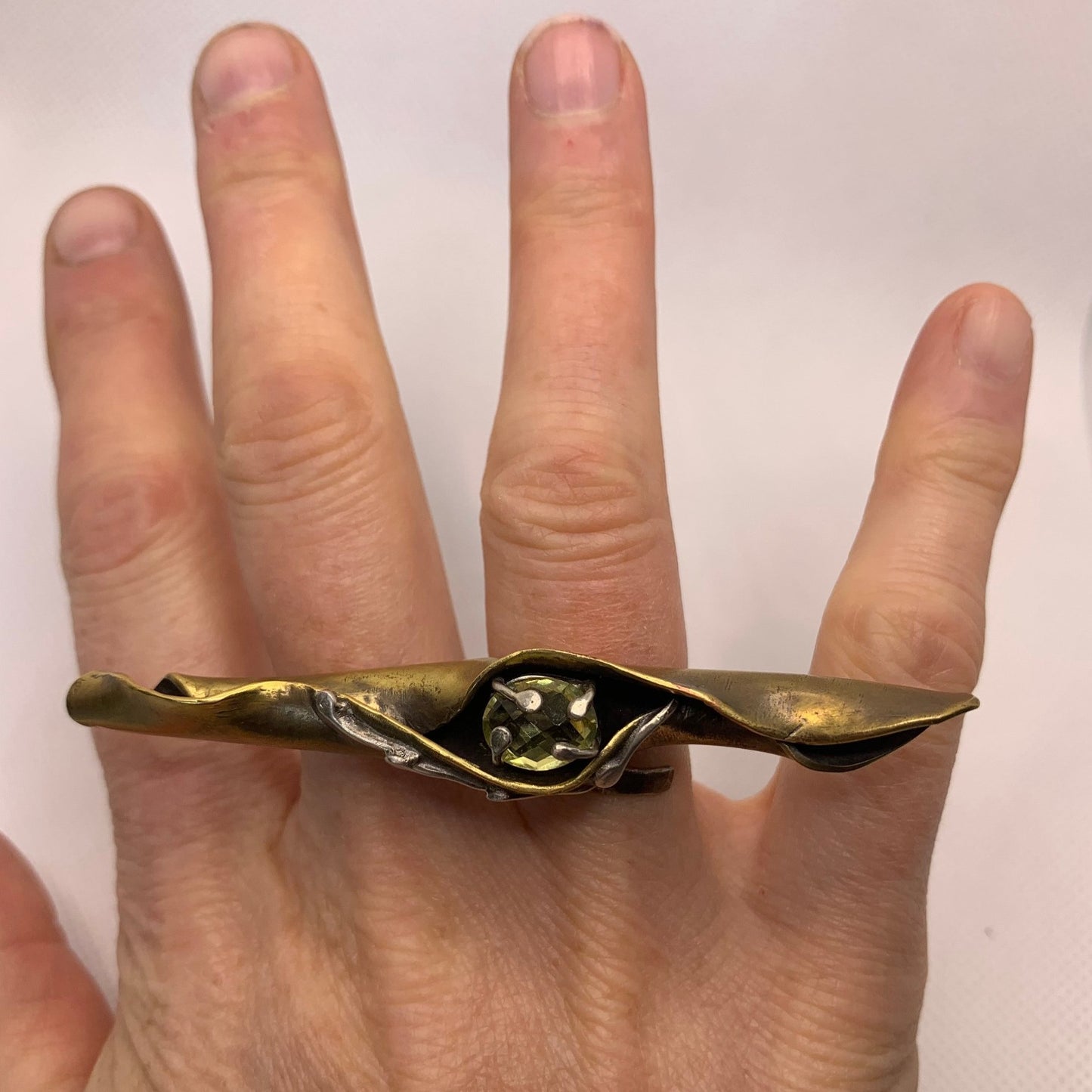 Bague en laiton forgé comme une feuille d'arbuste enroulée par le vent, elle couvre  4 doigts, repose sur un jonc réglable en laiton. En son cœur repose un serti en argent avec un quartz jaune facetté. Vue portée.